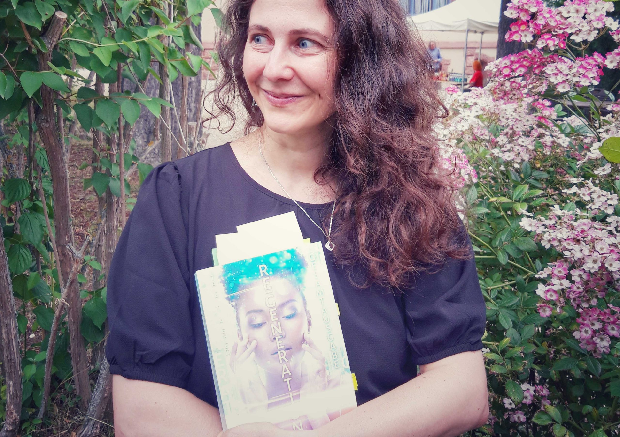 Tala T. Alsted steht vor einem blühenden Busch und hält ihr Buch "RE-GENERATION" in der Hand